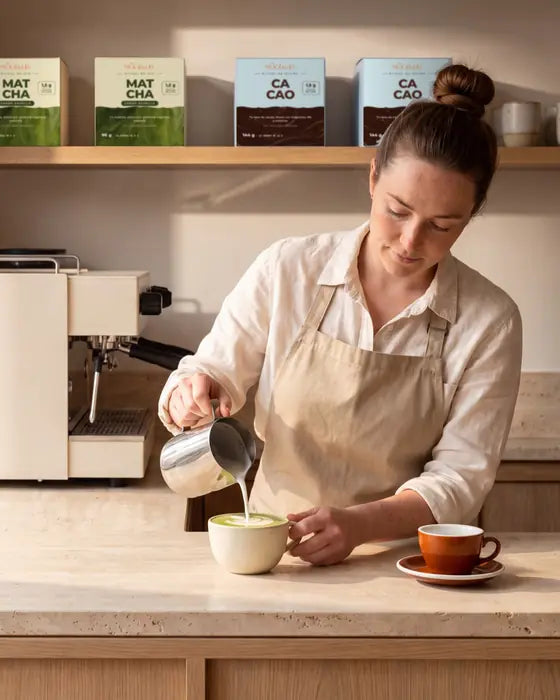 Barista vertiendo leche sobre un matcha en la barra de una cafetería, con cajas de Matcha Ritual y Cacao Ritual en la balda.