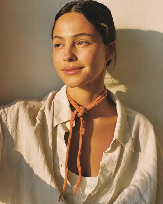 Mujer con camisa de lino y un cordón naranja al cuello, con los ojos entornados al sol de la mañana.