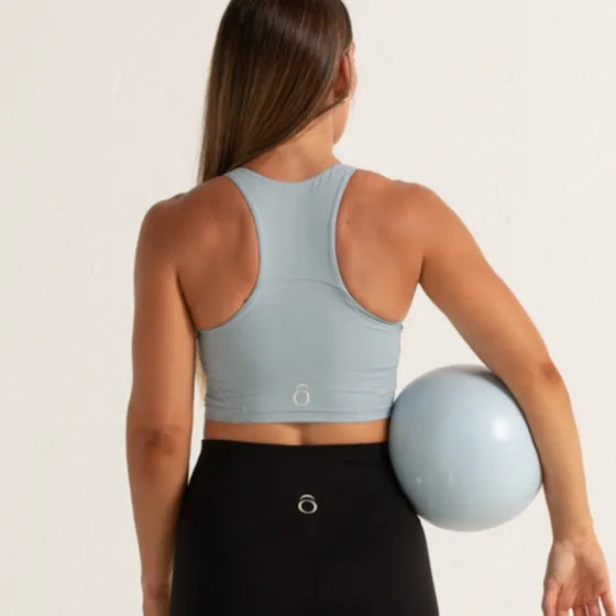 Top azul claro de espalda de nadadora visto por detrás, con legging negro y una pelota de pilates bajo el brazo.