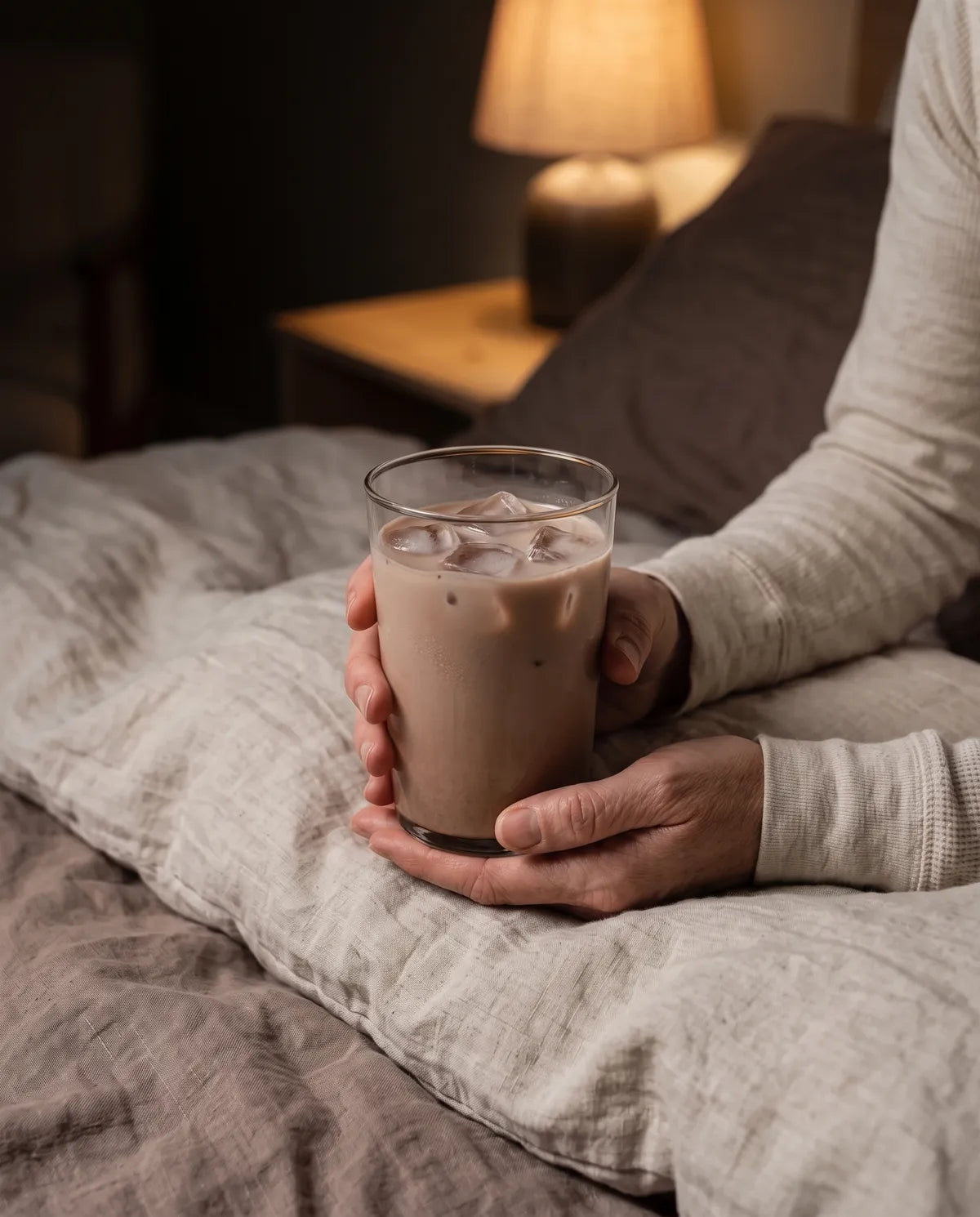 Unas manos sostienen un vaso de Cacao Ritual frío con hielo, sobre la cama, con una lámpara encendida al fondo.