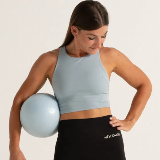 Mujer con crop top azul claro y legging negro, con una pelota de pilates bajo el brazo.