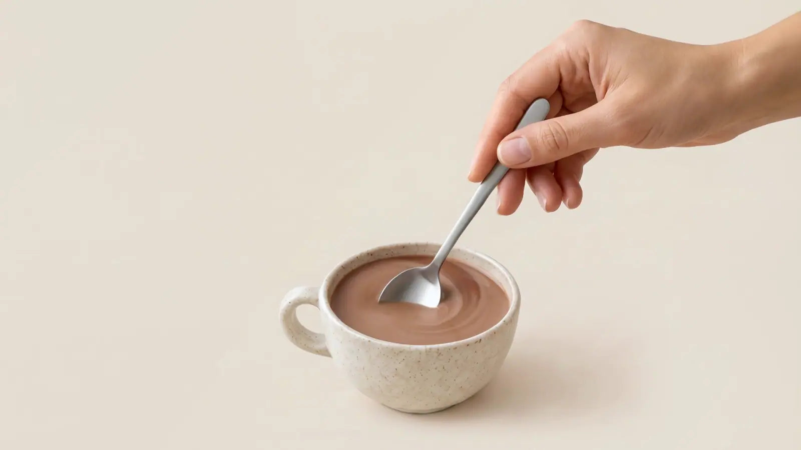Una mano remueve con una cucharilla la taza de Cacao Ritual hasta dejarla de un marrón cremoso.
