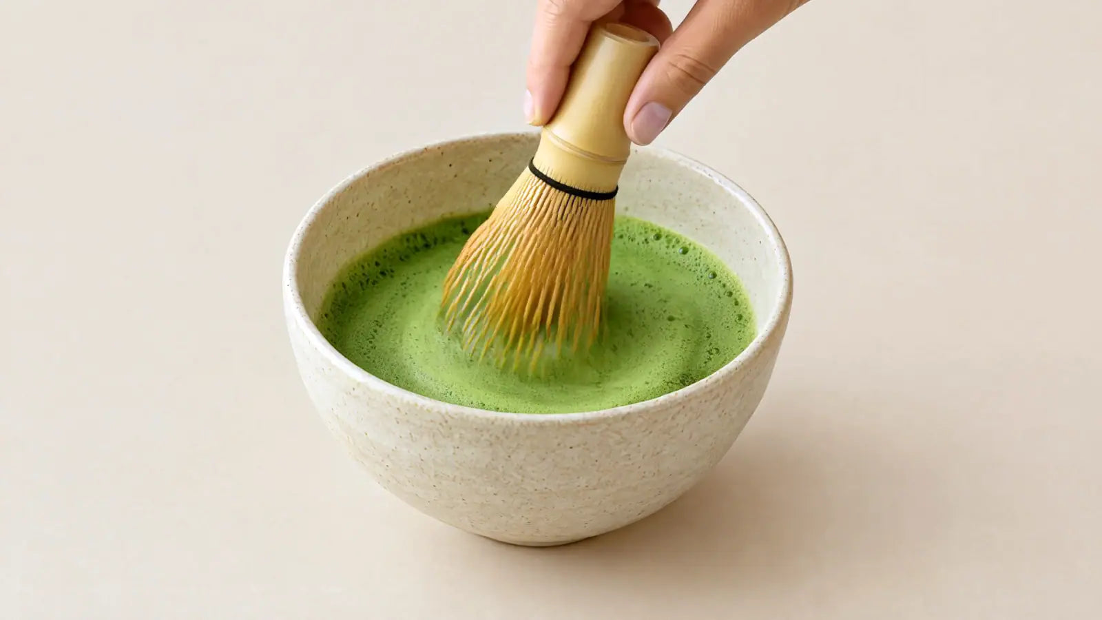 Una mano bate la bebida de matcha en el bol con un batidor de bambú hasta dejarla verde.
